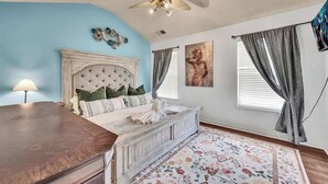 Un dormitorio con cabecera capitonada, cómoda de madera, alfombra floral, ventilador de techo y ventana con cortinas.