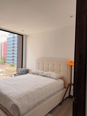 Ein Schlafzimmer mit Bett, Stuhl, Nachttisch und Fenster mit Blick auf Gebäude.
