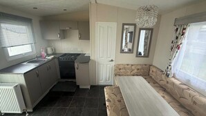 A compact kitchen with a stove, sink, and cabinets, a dining area with a bench, and a large window with curtains.