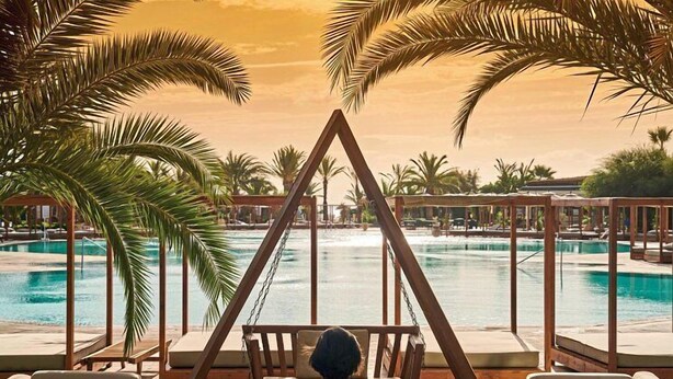 A person relaxing on a swing chair overlooking a pool area with palm trees and lounge chairs. - Sentido Marillia Hammamet  (Hammamet)
