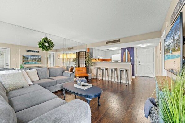 A living room with a grey sofa, a coffee table, and a kitchen area with bar stools. - MB Five Seasons 6203- 1BR Close to Beach (Myrtle Beach)