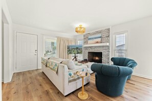 A living room with a sofa, a blue armchair, a fireplace, and a view of the city through a large window. - Endless Summer - Cherry Grove - 1 Block from Beach (North Myrtle Beach)