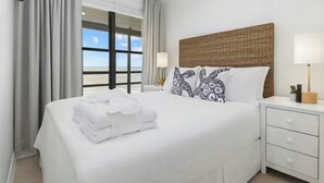 Ein Hotelzimmer mit einem großen Bett, zwei Nachttischen, einem Fenster mit Vorhängen und Blick auf den Strand.