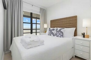 Ein Hotelzimmer mit einem großen Bett, zwei Nachttischen, einem Fenster mit Vorhängen und Blick auf den Strand.