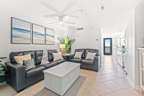 Ein modernes Wohnzimmer mit einem dunkelbraunen Ledersofa, einem weißen Couchtisch und einem Deckenventilator. An der Wand hängen drei gerahmte Kunstwerke, eine Topfpflanze, und es gibt einen Blick in den Küchenbereich.