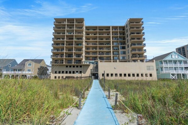 A multi-story building with balconies, a pathway leading to the entrance, and a beach in the background. - Oceanfront Buena Vista 703- 2BR- Outdoor Pool (North Myrtle Beach)