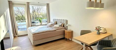 A modern bedroom with a bed, bedside tables, a dining area with chairs, and a view through large windows.
