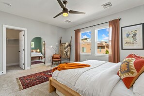A bedroom with a bed, a ceiling fan, a window with curtains, a chair, a mirror, and a door leading to another room.