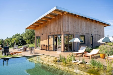 A modern wooden building with a pool, outdoor seating, and umbrellas.. Les Coteaux Lodges & Expériences