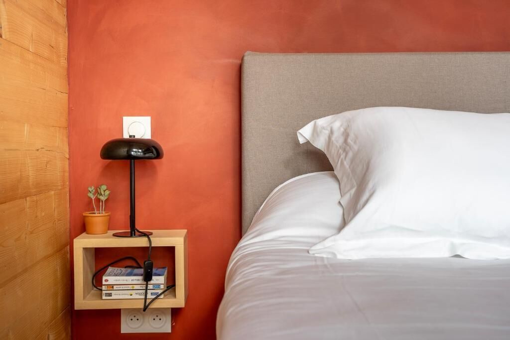 Una cama bien hecha con sábanas y almohadas blancas, una lámpara de noche, un pequeño estante de madera con libros y una planta, y un enchufe en la pared.