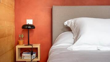 Una cama bien hecha con sábanas y almohadas blancas, una lámpara de noche, un pequeño estante de madera con libros y una planta, y un enchufe en la pared.