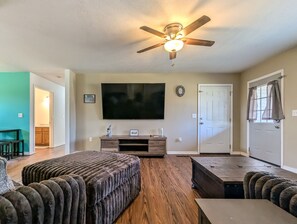 A living room with a ceiling fan, a flat-screen TV, a wooden entertainment unit, and a door with curtains. - Sugar Hill Home ~ simple & quiet 3BR near town (Mountain View)