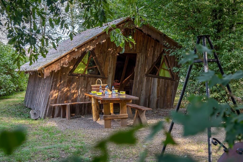 Una cabaña de madera rústica con una mesa de picnic y bancos, rodeada de árboles.