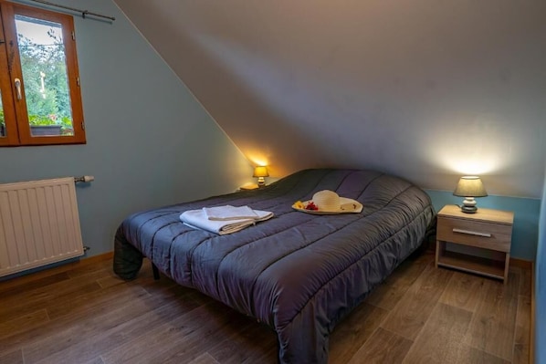 Un dormitorio con techo inclinado, una cama con cubrecama de color oscuro, dos lámparas de noche, una ventana con vista a follaje y un radiador.
