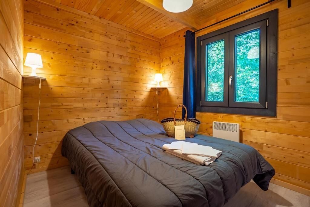 A wooden cabin room with a bed, a lamp, a basket, and a window.