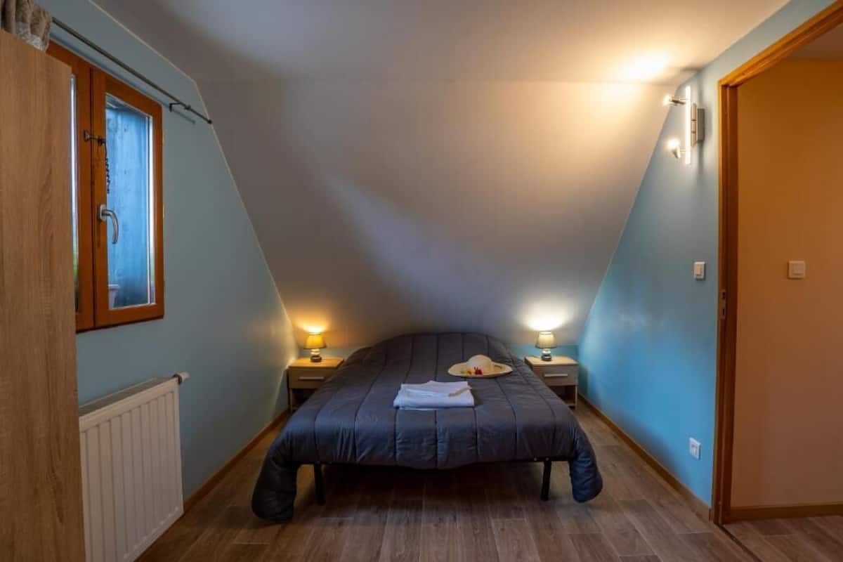 Ein Schlafzimmer mit schrägen Wänden, einem Bett mit dunkler Bettdecke, zwei Nachttischlampen, einem Fenster mit Vorhang und einem Heizkörper.