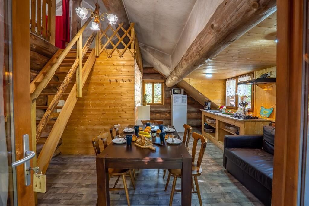 Un interior de madera con una mesa de comedor preparada para cuatro comensales, un sofá, una escalera y una zona de cocina.