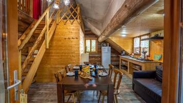 Un interior de madera con una mesa de comedor preparada para cuatro comensales, un sofá, una escalera y una zona de cocina.
