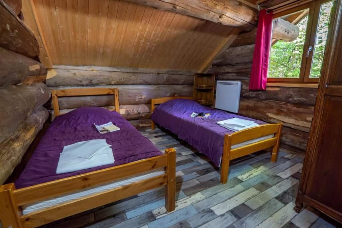 Ein Blockhauszimmer mit zwei Holzbetten, einem kleinen Fenster mit rotem Vorhang und einem weißen Heizgerät.