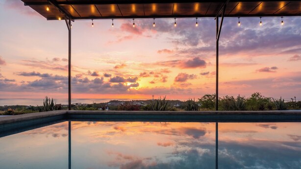 A swimming pool with a sunset view, a covered area with string lights, and a distant cross. - Casa Pola | Tropical Loft for 2 Near Beach & La Punta (Puerto Escondido)