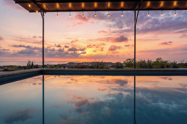 A swimming pool with a sunset view, a covered area with string lights, and a distant cross. - Casa Pola | Tropical Loft for 2 Near Beach & La Punta (Puerto Escondido)