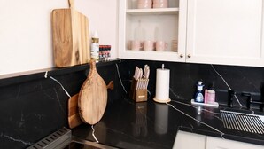 A modern kitchen with a black countertop, stainless steel range hood, wooden cutting boards, and a white cabinet with pink dishes. - Lil Pink Pool House • Coastal Cowgirl Charm • Walk to Historic Downtown McKinney (McKinney)