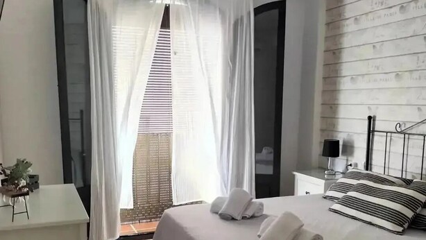 Un dormitori amb llit, una tauleta de nit, una làmpada, una finestra amb cortines transparents i vista exterior.