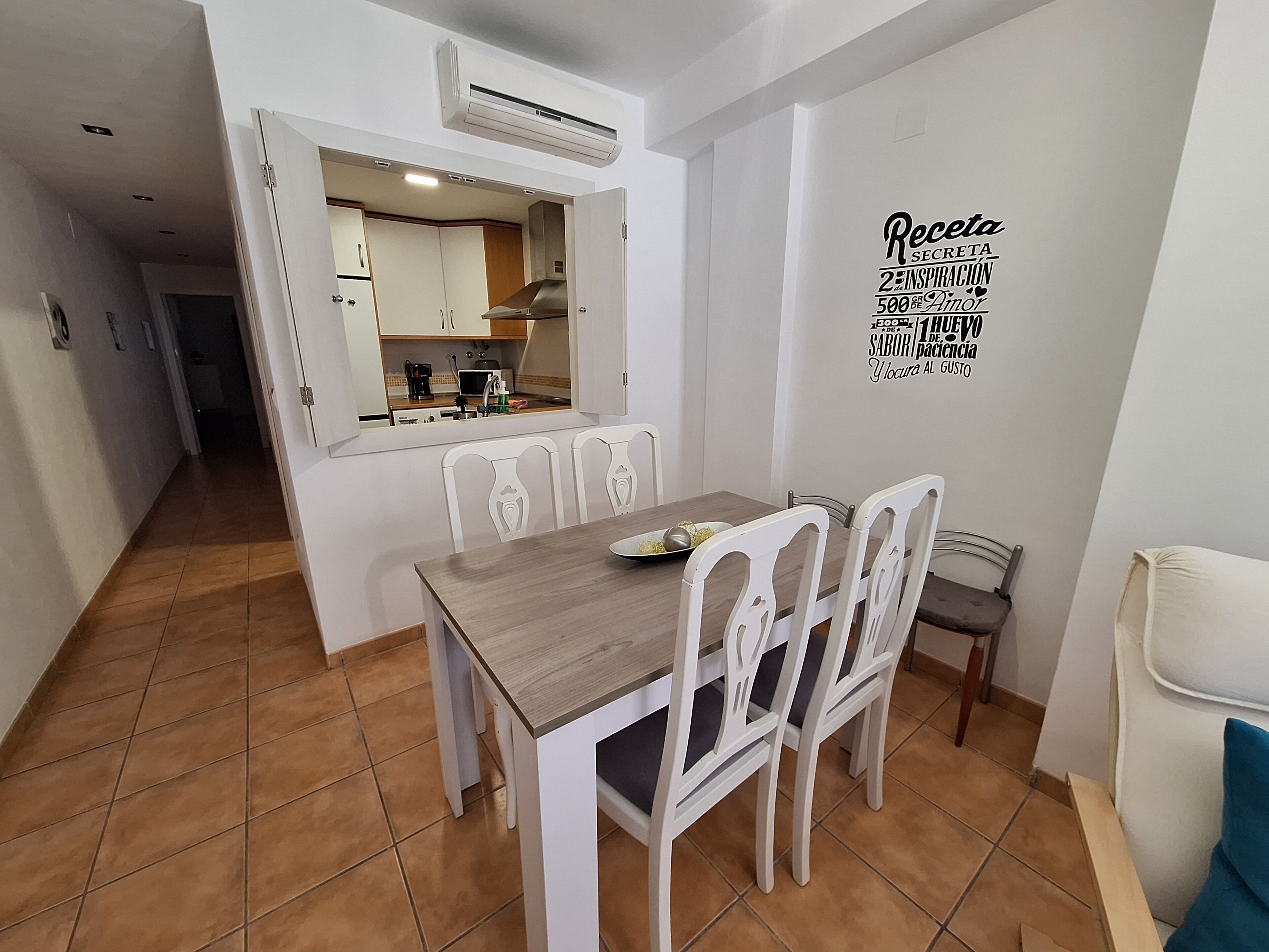 A dining area with a wooden table and white chairs, an air conditioning unit above, and a kitchen visible through an open door.
