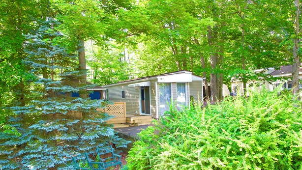 A mobile home with a porch and a small deck, surrounded by dense green foliage and trees. - Pocono Comfortable 2Bedroom Stay (Gouldsboro)