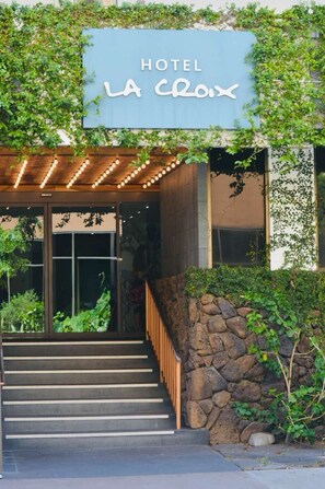 A hotel entrance with a sign reading 'HOTEL LA CLOIX,' a wooden staircase, and a stone wall.