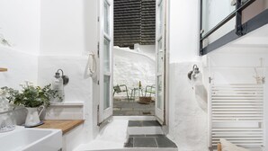 Un pasillo estrecho con una pared blanca, un lavamanos pequeño, una planta en maceta y una ventana con vistas a un patio.