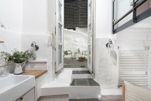 Un couloir étroit avec un mur blanc, un petit lavabo, une plante en pot et une fenêtre donnant sur un patio.