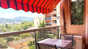 Un balcón con mesa y sillas, un toldo rojo y vistas a edificios y zonas verdes.