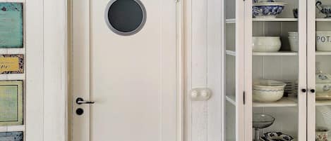 A white door with a round window, a cabinet with glass doors displaying dishes, and various decorative signs on the wall.
