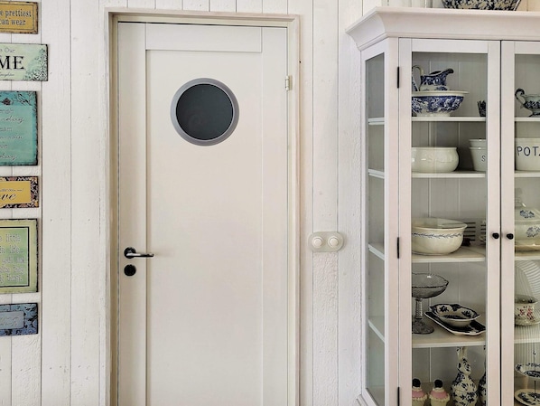 A white door with a round window, a cabinet with glass doors displaying dishes, and various decorative signs on the wall. - 5 Star Holiday Home in Ellos (Ellos)