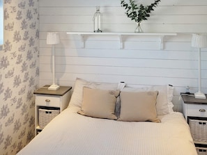 Un dormitorio con una cama, dos mesitas de noche, una lámpara, una estantería con un jarrón y una pared con estampado floral.