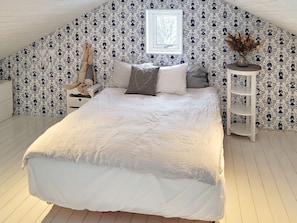 A bedroom with a sloped ceiling, patterned wallpaper, a bed with white bedding, a bedside table, and a small window. - 5 Star Holiday Home in Ellos (Ellos)