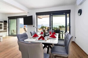 A dining area with a table set for four, a TV mounted on the wall, and a view of the outdoors. - Holiday house LAVANDA with pool and panoramic seaview (Risika)