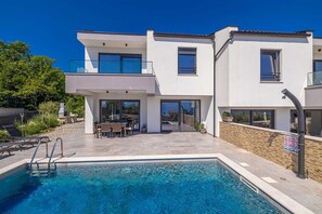 Una casa moderna de dos pisos con piscina, mobiliario exterior y un muro de piedra.