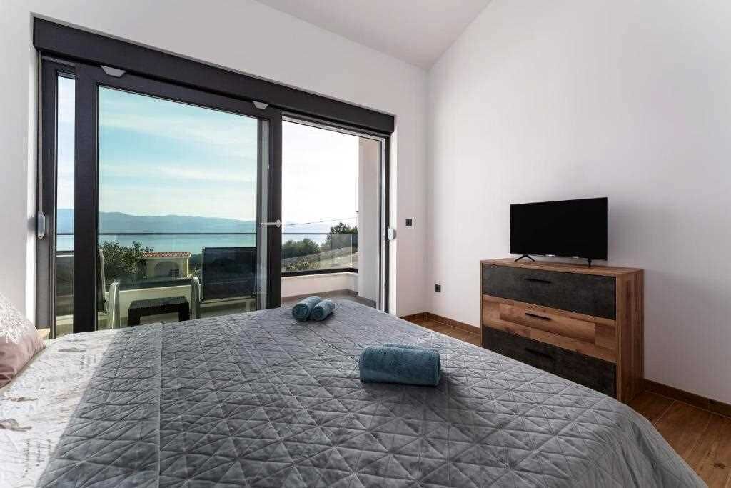 A bedroom with a bed, a TV on a wooden dresser, and a sliding glass door leading to a balcony with a view.