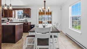 A modern kitchen with a central island, wooden cabinets, a dining table with chairs, and a large window with a view. - Spacious 4BR Home Sleeps 10 I Game Room + Sunroom+ Veranda (DIEPPE)