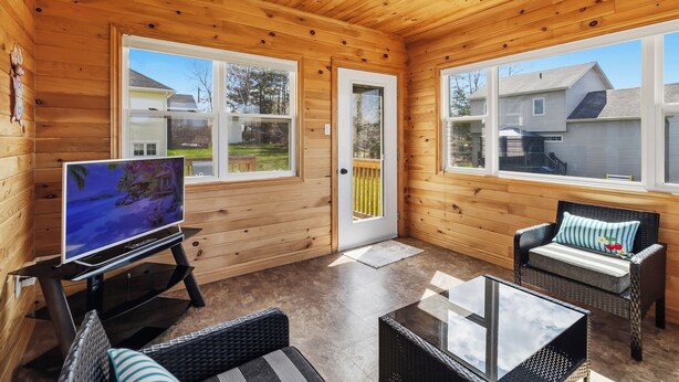 A wooden cabin with a TV, wicker furniture, and large windows. - Spacious 4BR Home Sleeps 10 I Game Room + Sunroom+ Veranda (DIEPPE)