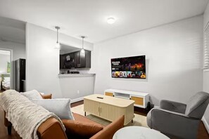 Ein modernes Wohnzimmer mit Sofa, Sessel und Couchtisch. Eine Küchenzeile mit Schränken und Kühlschrank ist ebenfalls vorhanden. Ein Flachbildfernseher ist an der Wand befestigt.