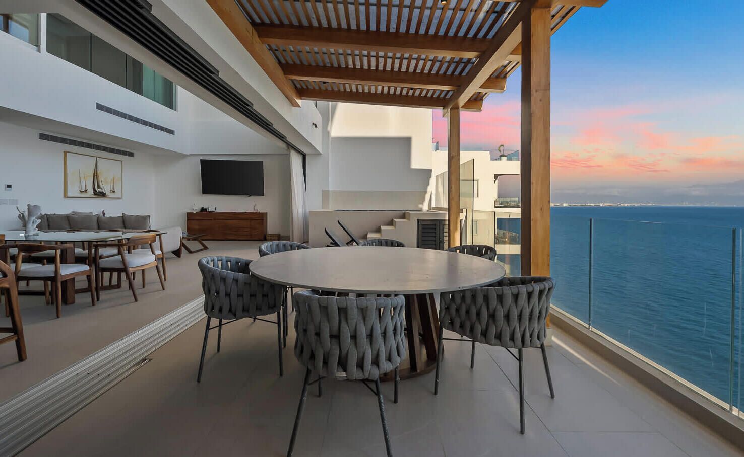 A modern outdoor dining area with a round table and chairs, a sofa, and a TV on a white wall.