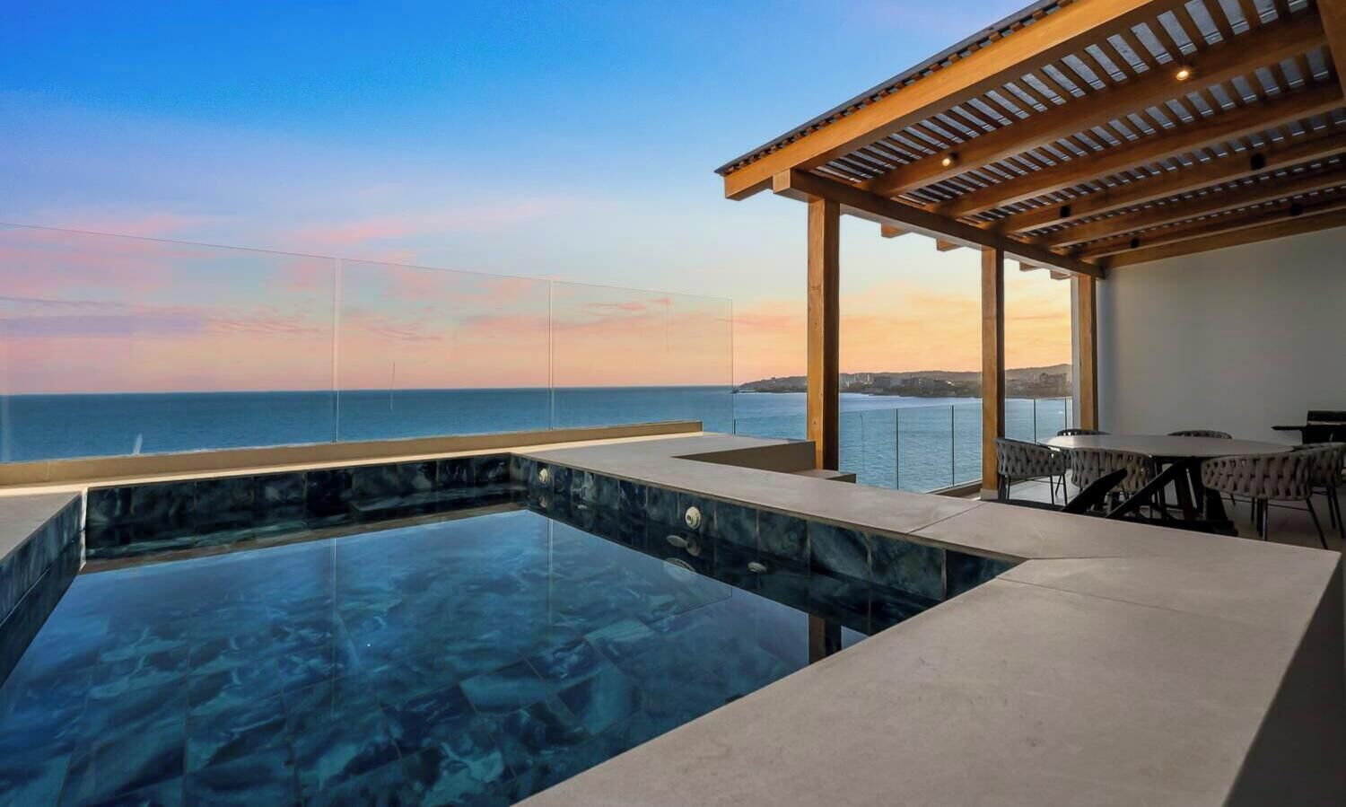A modern infinity pool with a clear view of the ocean, a covered outdoor seating area, and a dining table.