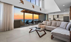 A modern living room with a large sofa, a coffee table, and a view of the ocean at sunset. - Lux Beachfront 5BR Penthouse Retreat | Panarea (Bucerías)