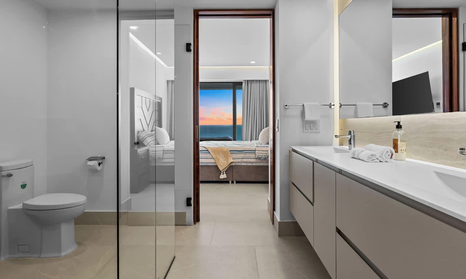Ein modernes Badezimmer mit Toilette, einer Glasabtrennung, einem Waschbecken mit Spiegel und Blick auf ein Schlafzimmer mit Bett und Vorhängen.