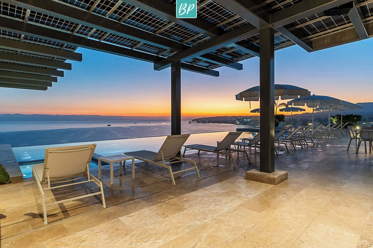 Une terrasse sur le toit offrant une vue sur l’océan au coucher du soleil, avec des chaises longues, une piscine et un espace abrité doté de parasols.