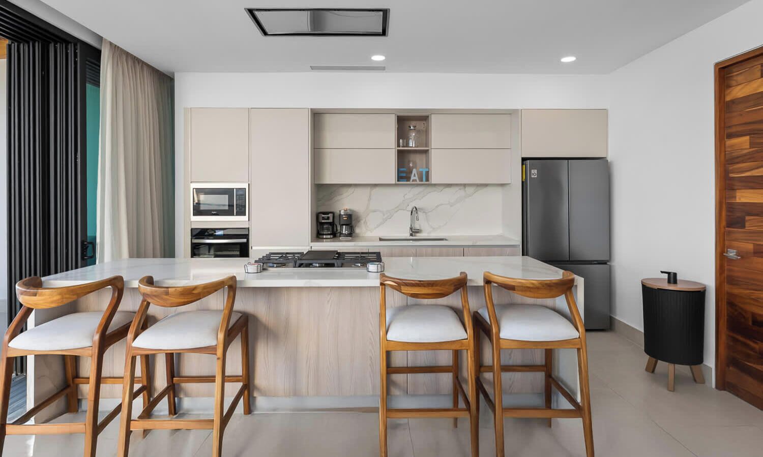 A modern kitchen with a central island, bar stools, a refrigerator, and a coffee machine.