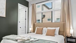 Una cama bien hecha con una manta verde, cabecera blanca y cortinas color beige.